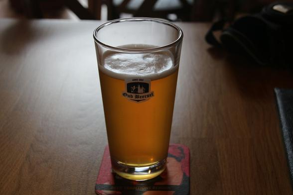 A straight sided glass of hazy golden beer. There is very little froth. The glass is printed with the Oud Beersel logo, which shows the outline of Beersel castle.