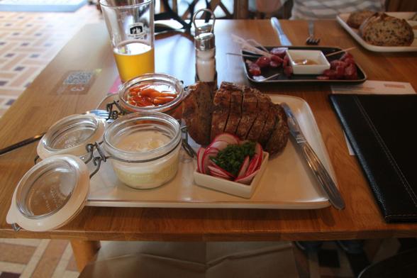 A glass pot of curd cheese, a glass pot of pickled vegetables, a small container of thinly sliced radishes, and a sliced half loaf of rustic brown bread. All arranged on a white oblong plate.