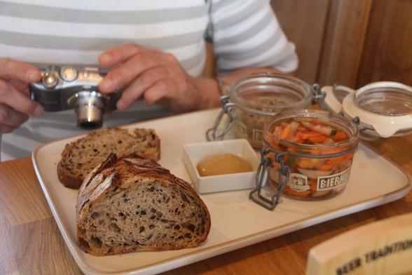 A glass pot of coarse paté, a glass pot of pickled vegetables, a small container of mustard, and a sliced half loaf of rustic brown bread. All arranged on a white oblong plate. My partner Sarah is holding her camera in the background.