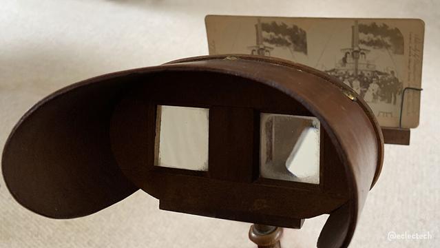 A photo of an old wooden stereoscope, with a stereoscopic image in the holder. The stereoscope has a lens for each eye with a wooden surround to stop peripheral view / light and a barrier to separate the view for each eye. Beyond the headset is a holder for stereoscopic images (two images depicting left-eye and right-eye views of the same scene). Viewing through the device means that for most people they are shown as a single three-dimensional image. The image in this photo is a group of people on a boat. It is a thing of beauty and charm.