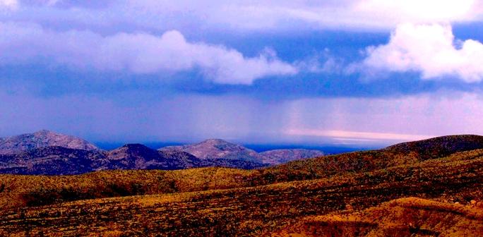 A desert landscape featuring a distance rainstorm. In the foreground are short, orange hills without vegetation. Behind that are taller hills that appear blue, and are also without vegetation. Behind that is a blue cloudy sky, with two curtains of rain reaching the landscape.