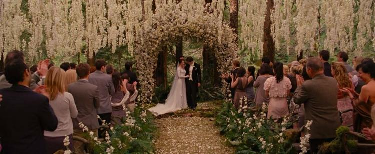 A bride and groom stand under a huge pink floral arch as their guests all stand and applaud
