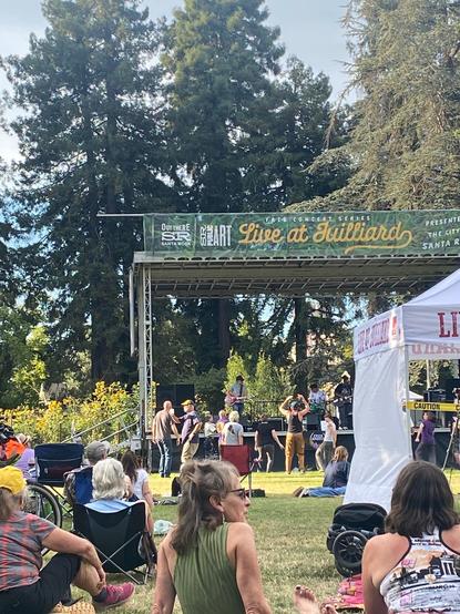 Outdoor stage surrounded by redwood with sign that says live at Juilliard. People sitting and dancing in front of stage.
