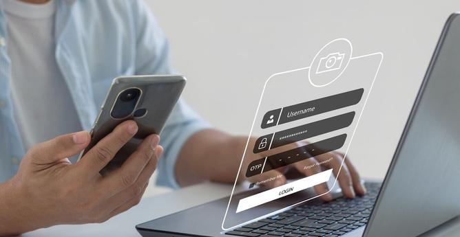 Image of a man working on his laptop with his mobile phone, where the login screen renders over the keyboard.