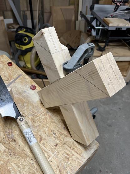 A wooden X is clamped to a workbench. Now from the other side. There are markings on the ends of the X designating where it has to be cut.