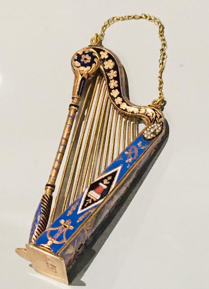 Photographie en couleur d’une minuscule harpe en or et émail, d’environ 7 centimètres. 
La caisse de résonance qui dissimule la boîte à musique est ornée de délicats motifs floraux et musicaux sur un fond bleu émaillé. 
Une fine chaînette de suspension en or est fixée sur le haut de la harpe.