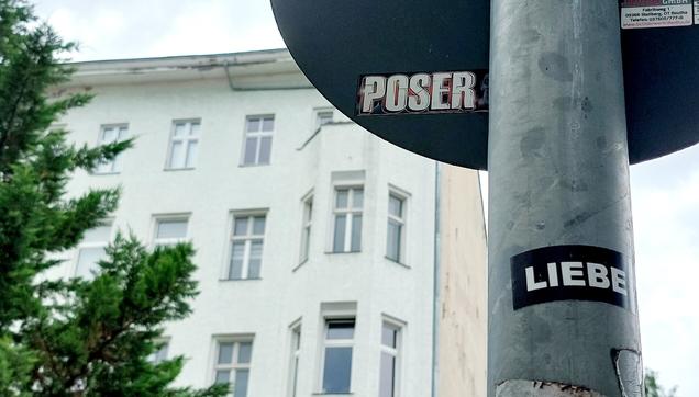 Im Hintergrund ein Berliner Altbau und ein Baum. Im Vordergrund ein Metallpfahl und die untere Hälfte eines rundes Straßenschildes. Auf dem Schild ein Aufkleber mit dem Wort POSER, auf dem Pfahl ein Aufkleber mit dem Wort LIEBE.