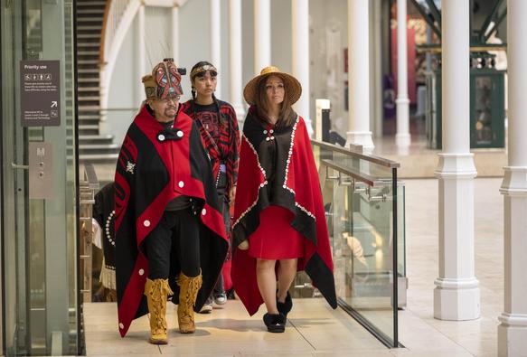 Three people walk in a building. They are dressed in West Coast First Nations regalia. The man wears a cedar carved headdress. One woman wears a cedar woven hat and the other woman a cedar headband with abalone shell as an adornment. They wear red and black button blanket shawls.