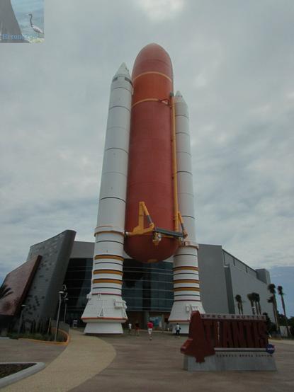 An outdoor photograph of the facade of the Kennedy Space Center Visitors Complex’s Atlantis Museum, which is black and angular, with a full-sized replicas of the Space Shuttle’s orange External Tank and white Solid Rocket Boosters.