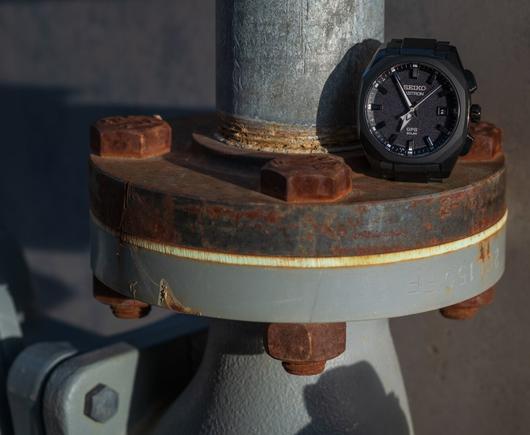 Some outdoor metal plumbing-y bits, in hard sunlight.  Some parts are painted gray, some are attractively rusted metal.  There is a black wristwatch propped atop a rusted metal disc, leaning against a silvery anodized-looking vertical pipe.