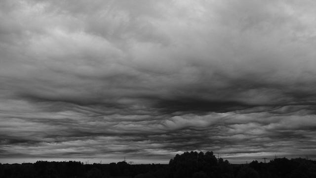 Schwarzweißfoto einer dramatisch wirkenden Wolkenformation über der Silhouette einer Landschaft mit Bäumen, Strommasten und Windrädern.