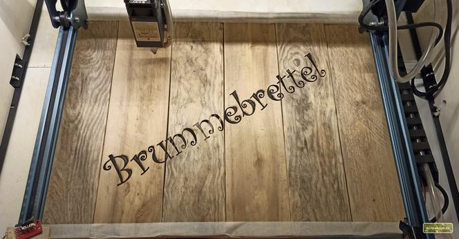 mehrere alte Holzbretter liegen in einem CNC-Laser. Diagonal wurde "Brummebrettel" in einer verschnörkelten Schriftart eingraviert.
