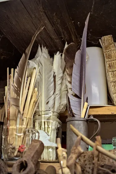 Verschiedene Dinge in einem Regal. Viele Vogelfedern, kleine Schnitzereien, Papier. Im oberen Bereich ein Holzbalken, viele Spinnenweben.
---
Various things on a shelf. Many bird feathers, small carvings, paper. In the upper part a wooden beam, many spider webs.