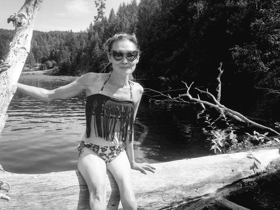 Monochrome photo.
Me, in a fringe top bikini, sitting on driftwood log that is floating on a lake. Wearing heart shaped sunglasses.