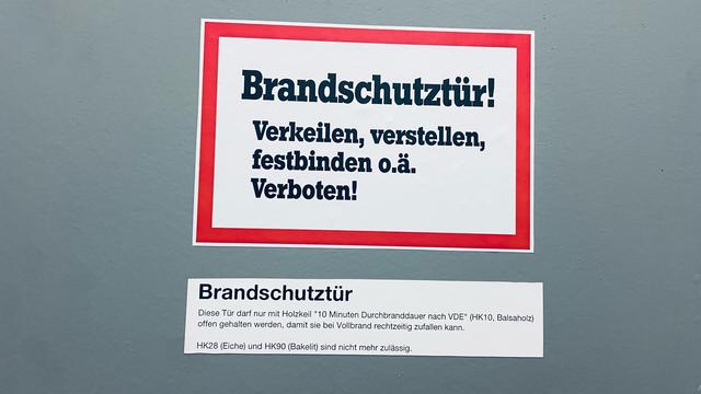 Zwei Aufkleber: 1) „Brandschutztür! Verkeilen, verstellen, festbinden oder ähnlich verboten!“ 2) „Brandschutztür. Diese Tür darf nur mit Holzkeil 10 Minuten Durchbranddauer nach VDE (HK10 Balsaholz) offen gehalten werden, damit sie bei Vollbrand rechtzeitig zufallen kann. HK28 (Eiche) und HK90 (Bakelit) sind nicht mehr zulässig“