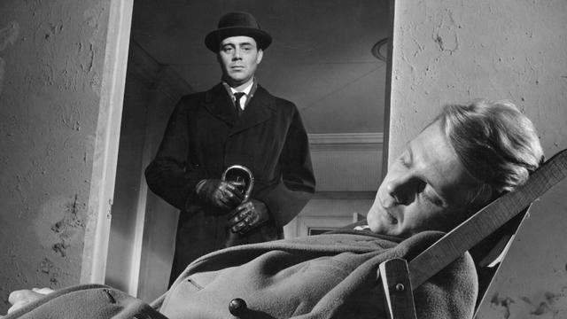 Manservant Barrett (Dirk Bogarde) and his master Tony (James Fox). Tony is asleep in a folding chair wearing an overcoat in a sparse room with crumbling walls. Barrett – immecuatly dressed in a dark overcoat, hat, gloves, tie – stands in the door opening, looking down on the sleeping man.