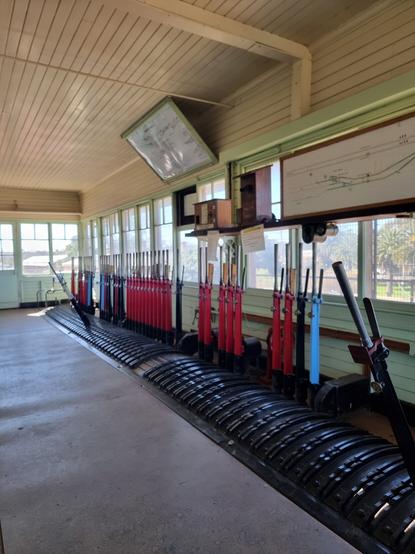 The interior of a 1880s era signalbox, showing the levers that control signals (red) points (black) and locking bars (blue).  Most of the levers are in the normal (upright) position but there is one in the foreground and one towards the far end that are pulled. The lever frame sits in a large room with windows all around through which the signaller can see the movements of the trains around the station