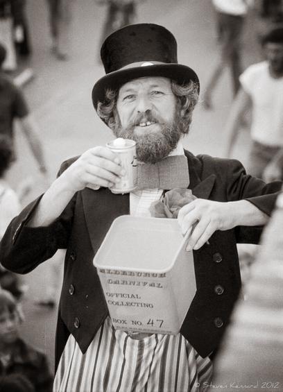 A black and white photo of a bearded man in a top hat who is on stilts, so high above the people walking below him. He is smiling in a friendly way and holding a can of beer with froth on the top and a square box with "Aldeburgh Carnival" printed on the side, for donations.