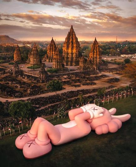 Photography and exhibition. A color photo of a 45 meter long artwork lying on a lawn in front of a temple complex in Indonesia. The figure is a pink creature with wavy ears and white Mickey Mouse gloves. It is lying in the grass with its hands over its eyes.
Info: The figure is part of the world exhibition tour of the American artist "Kaws", which began in 2018. On its last stop, the exhibition has landed in Indonesia: 19-31 Aug 2023 Hours: 8am-5pm (There is a fee to enter the Prambanan Temple Park Complex).
