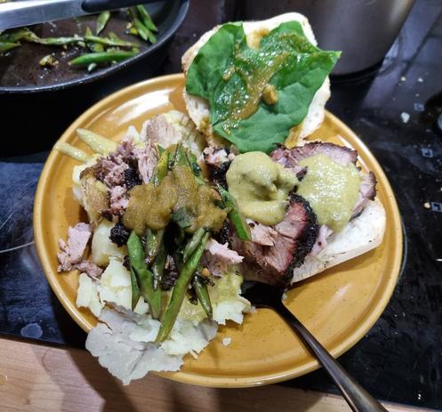 A shallow orange bowl of food. Pork in a bread roll and on crushed potatoes, with a pale green salsa on and some cooked beans and green chilli sauce, there are some token leaves on the top half of the roll.