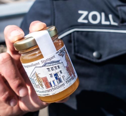 Ein Zöllner in Dienstkleidung hält ein Glas Sommerblütenhonig in der Hand, der auf dem Flachdach des Hauptzollamts Stuttgart von vier Bienenvölkern produziert wird.