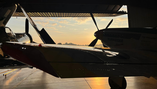 Color image of P-51 Mustang and Corsair FG-1D WWII-era aircraft poised in the hanger and ready for the airshow. Image shows the Corsair in blue, with the wings folded up, the Mustang in silver and red just behind the Corsair, and the hanger door open to the rising sun, fluffy clouds in a blue sky above.