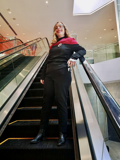 Jil laughs as she rides up the escalator in her Captain Seven of Nine Cosplay. She’s wearing a red & black Starfleet uniform and is wearing silver Borg prosthetics on her hand and face.
