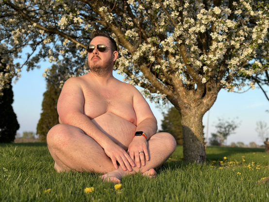Sky-clad man seated cross-legged under a blossoming crab apple tree in the bright sunlight.