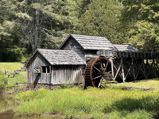 An old, restored water mill on a stone foundation with a large water wheel on the side. The building is made of weathered wood boards which have turned almost grey, and matching grey shingles. .