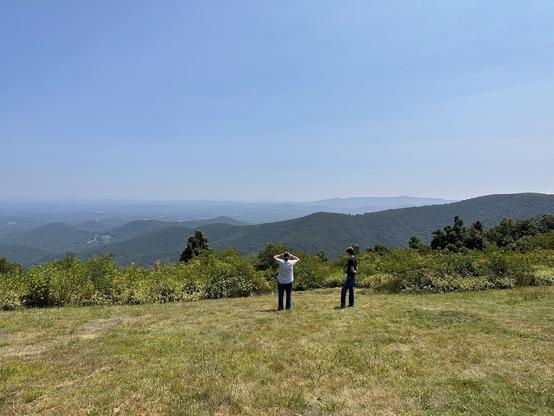 A woman and a teenage boy standing in a field, facing away from the camera, looking out at the Appalachian mountains. Several mountain ridges are visible in the distance, until atmospheric haze obscures the rest of the view.