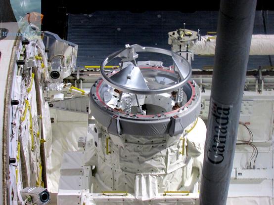 An indoor photograph of a cylindrical tunnel inside the cargo bay of the Space Shuttle, with a gray top with three petals on top.