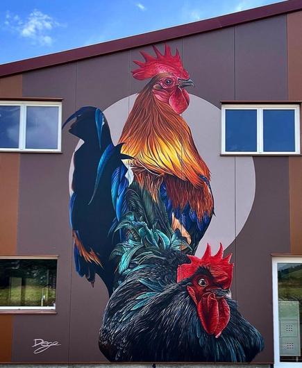 Streetartwall. On the outer wall of a modern one-story house with 3 windows and a glass door is sprayed/painted a mural of a huge rooster with a chicken. On brown background is sprayed a magnificent rooster with yellow and royal blue plumage. In front of him sits a green black chicken.