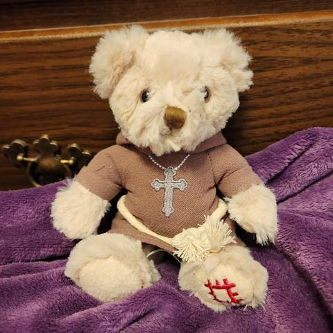 A small teddy bear wearing brown monk's robes with a cross embroidered on it with a rope belt, sitting on a purple blanket.