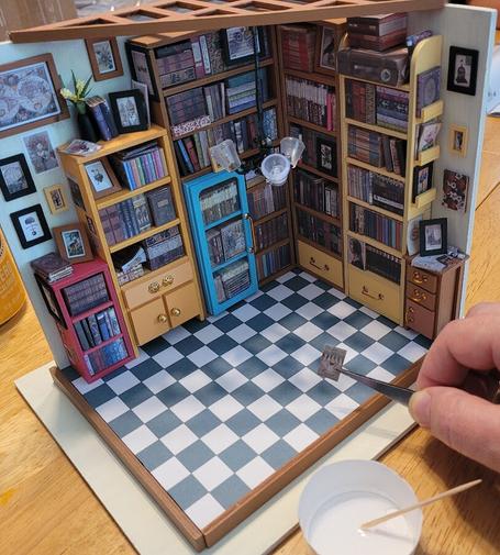 A miniature diorama of a home library with two walls populated with bookshelves and framed pictures. A human hand with tweezers holding a tiny picture is creeping into the scene. A truncated cup of glue and toothpick remind us that this is all glued together, much like reality.