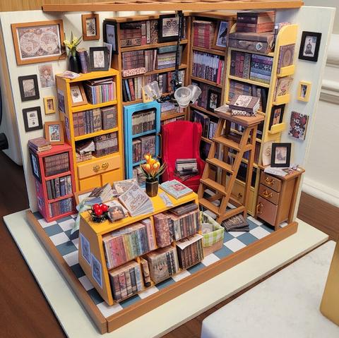 The completed library diorama with framed pictures, paper flowers in vases, books and papers. An armchair, ladder, and low angled bookcase have been added to the scene. There are books everywhere. It is bright and cheery. I want to be in there.