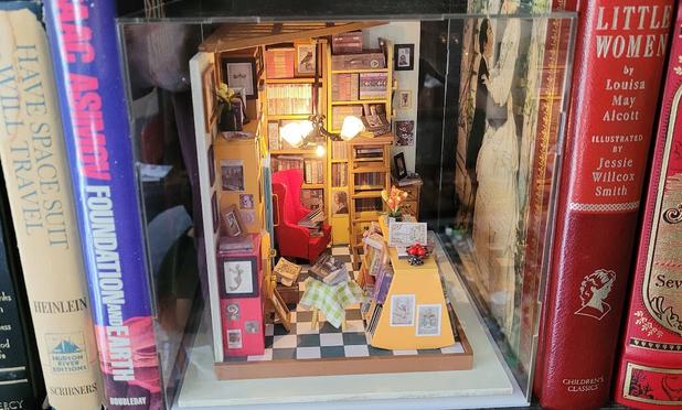 The diorama is in a clear acrylic case and sits on a real bookshelf between real, full-sized books. The LED chandelier is lit and the diorama shines warm light onto itself.