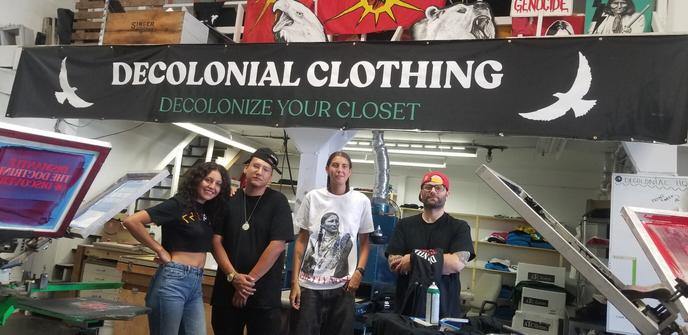 The all Indigenous crew at Decolonial Clothing Co warehouse shop in Burnaby, BC. There are screen printing machines on either side, framing the crew. Their business banner hangs above & text reads:
Decolonial Clothing
Decolonize Your Closet