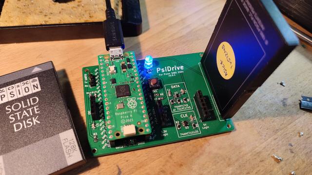 A photo of the PsiDrive, fully constructed with a Raspberry Pi Pico on the top. It has a Spoon SSD plugged into it. To the left is a pile of SSDs.