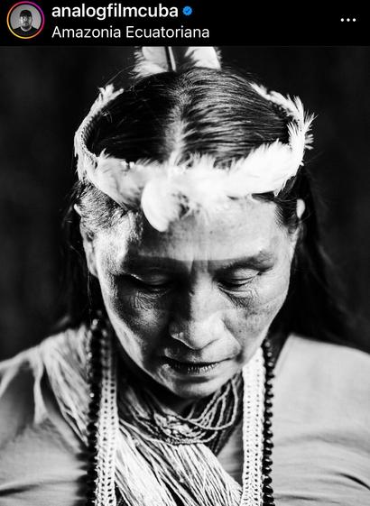 Black and white image of indigenous woman looking down She is wearing many strands of beads and has a crown of white feathers.