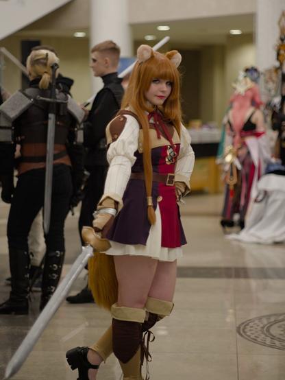 A girl wearing short white dress with long sleeves. On top of the dress she wears brown and red camisole stylized to medieval one and brown shoulder straps. Her right hand is lowered, she holds a sword pointing down and to the right of her. Her left hand is on her hip. She wears red-brown wig with raccoon ears, behind her legs raccoon tail is visible.