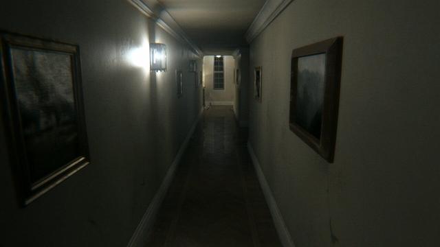 Pasillo de una casa muy largo únicamente iluminado por una bombilla que no llega a cubrir todo el pasillo. En el fondo, una ventana que muestra el exterior nocturno, y el pasillo sigue a la derecha. El suelo de madera oscura, y las paredes grises, con rodapiés y cornisas blancos. En las paredes, cuadros con marcos grandes de madera.