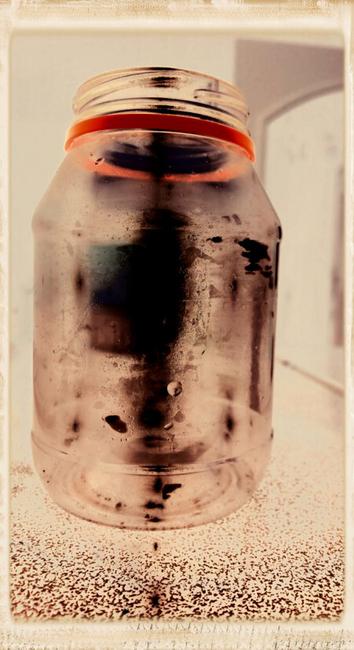 The composition is a faux antique photograph with albumen print edge. The subject is a medium close-up of an empty jar without a lid. The tones are beige and reddish browns. There is a surreal atmosphere, the jar has a dark ghostly glow.