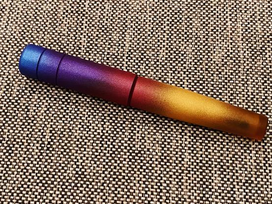 A fountain pen by the Filipino brand Kasama, called the Una Takipsilim. It has a vivid gradient from metallic blue, to purple, red, and gold, fading at the end to show off the raw Ultem material the pen is made of (a translucent yellow plastic material that’s very strong). The pen is sitting on a gray tweed couch cushion.