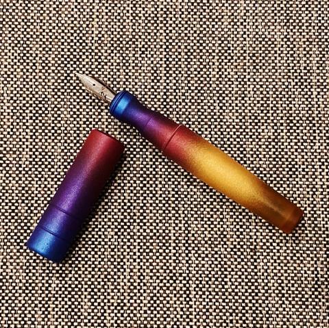 A fountain pen by the Filipino brand Kasama, called the Una Takipsilim. It has a vivid gradient from metallic blue, to purple, red, and gold, fading at the end to show off the raw Ultem material the pen is made of (a translucent yellow plastic material that’s very strong). The pen is sitting uncapped on a gray tweed couch cushion.
