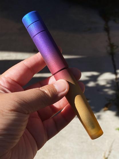 A fountain pen by the Filipino brand Kasama, called the Una Takipsilim. It has a vivid gradient from metallic blue, to purple, red, and gold, fading at the end to show off the raw Ultem material the pen is made of (a translucent yellow plastic material that’s very strong). The pen is held in bright, direct sunlight to show off its colors.