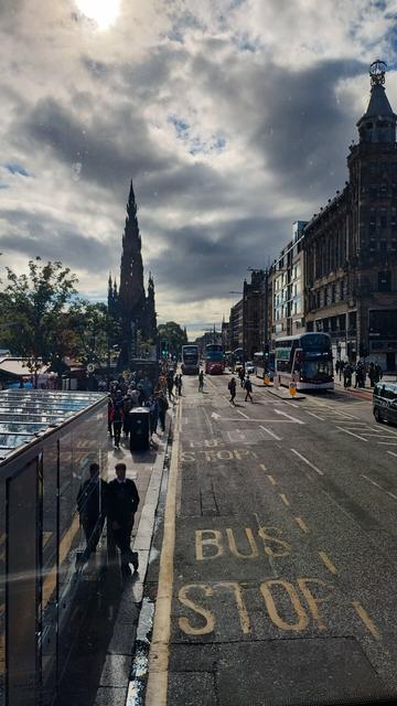 View from top deck of a bus, looking right along Princes Street as a mix of clouds and sun create unusual light quality, the Gothic rocket of the Scott Monument standing against the skies