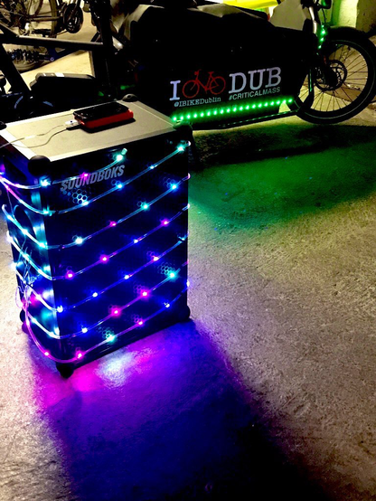 A music speaker wrapped with glowing lights. In the background is a Cargo Bike also donning glowing lights.