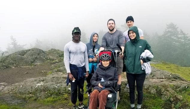 Me, sitting in trail rider chair, with my Power To Be support team. Foggy background, we are atop a small mountain summit.