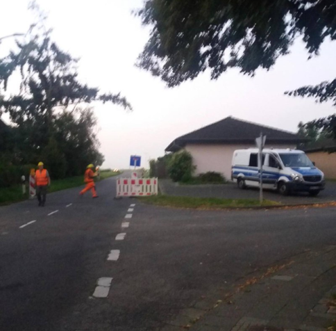 Bild aus dem L12 muss bleiben Ticker... 2 orange RWE Arbeiter*innen bei Absperrbake am Ortsausgang Keyenberg. Daneben Polizeisprinter auf Hauseinfahrt.