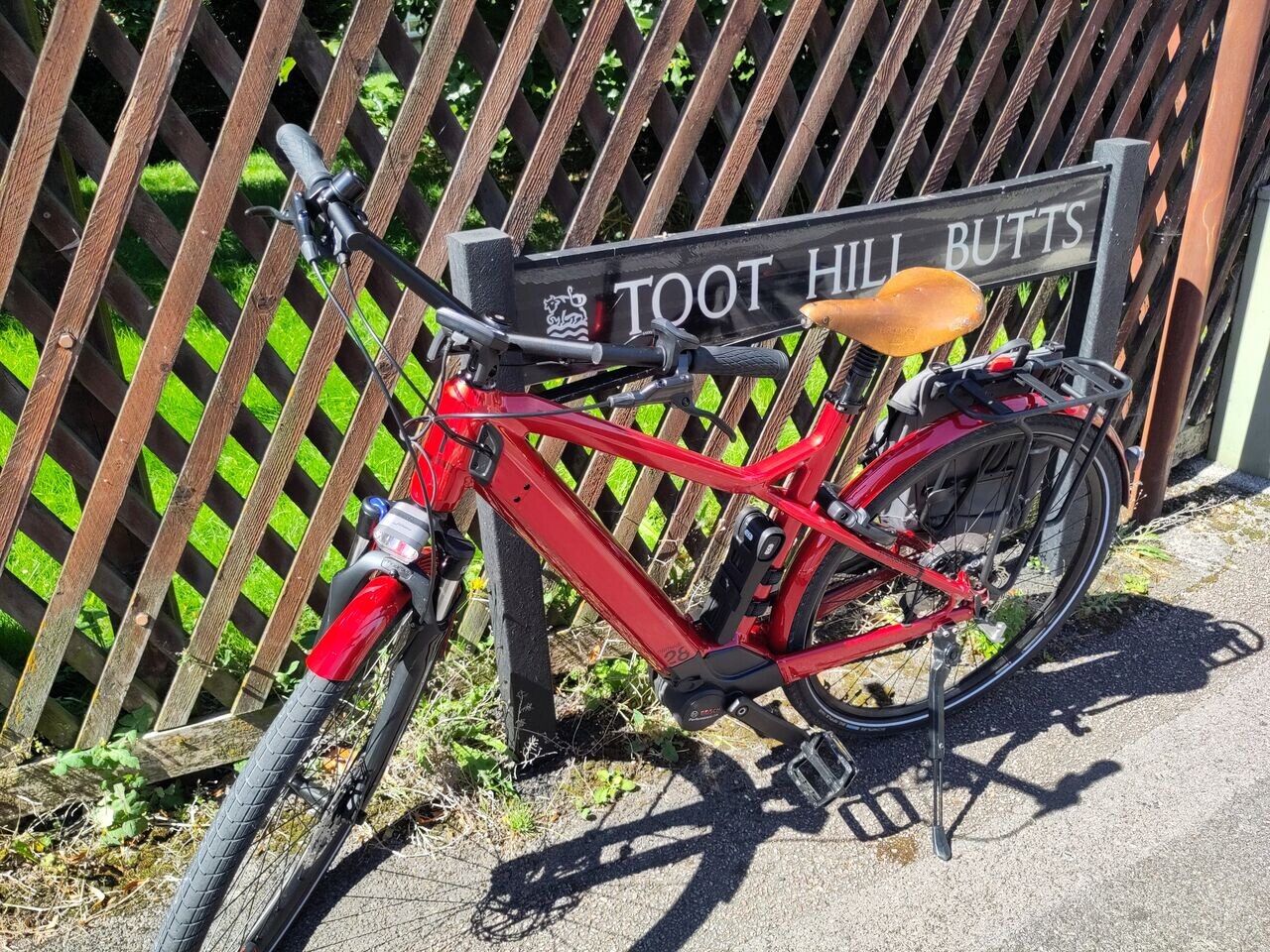 A red Moustache Samedi 28.5 electric bicycle next to a street name sign reading "Toot Hill Butts"